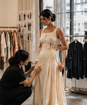 A fitter kneels to adjust an ivory dress in a rail-lined atelier
