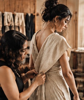 A fitter gathers a linen drape at a woman's waist in a wood-panelled studio
